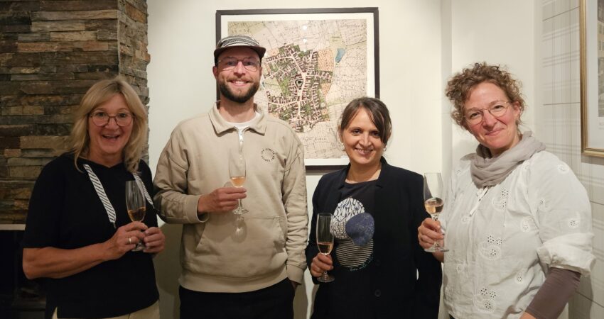 Heike Spitzner, Lukas Fassnacht, Ingrid Gutjahr und Christina Siebert lächeln in die Kamera. Sie halten jeweils ein Glas Sekt in der Hand. Im Hintergrund eine historische Karte von Gochsheim.