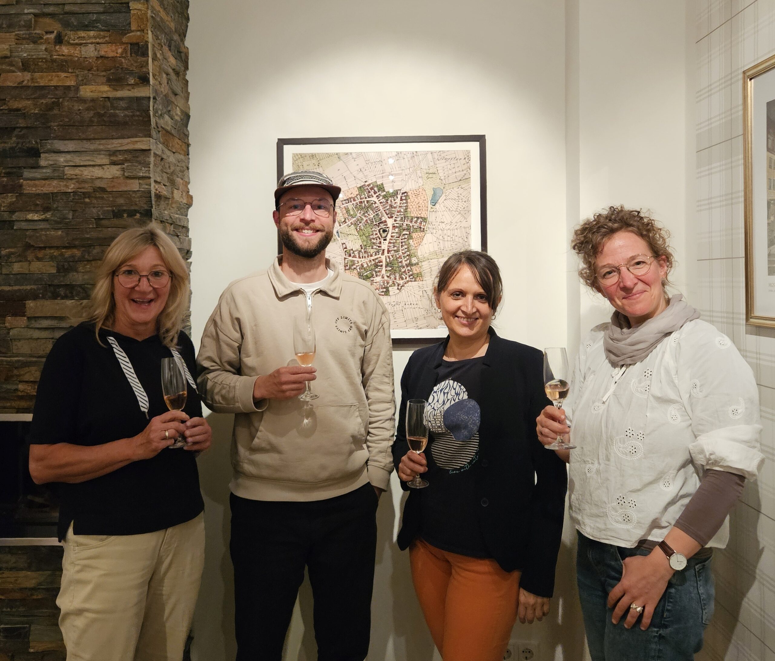 Heike Spitzner, Lukas Fassnacht, Ingrid Gutjahr und Christina Siebert lächeln in die Kamera. Sie halten jeweils ein Glas Sekt in der Hand. Im Hintergrund eine historische Karte von Gochsheim.