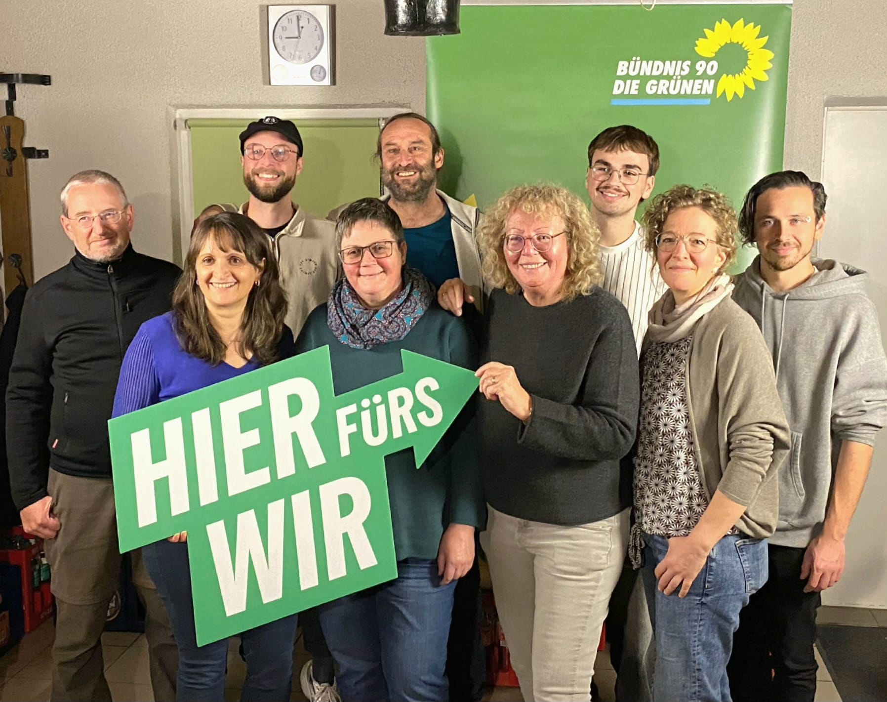 Gruppenfoto, im Hintergrund ein Banner mit dem Logo von Bündnis 90/Die Grünen. Die vier Frauen stehen im Vordergrund und halten ein großes Logo des Wahlkampf-Slogans "hier fürs wir" in Pfeilform.
